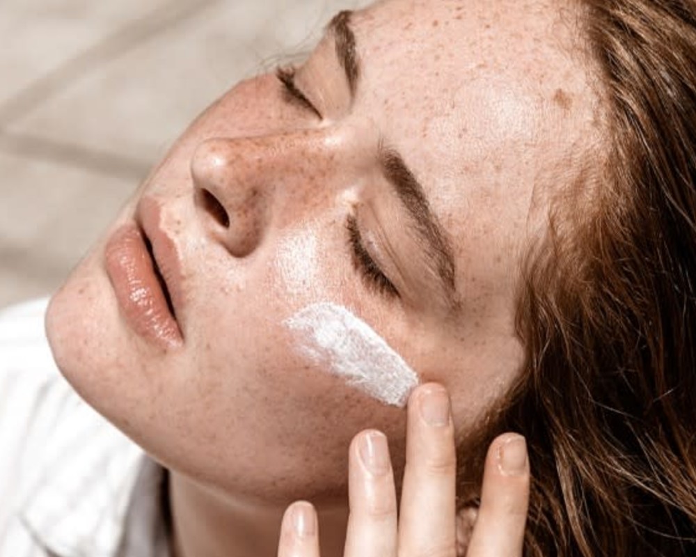 Ultimate Guide to Sunscreen - Woman with red hair and freckles. Sun is shining on her face, her eyes are closed and she is swiping sunscreen onto her cheek.