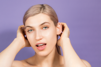 Adore Beauty - young woman with blonde hair is looking happy as she tucks hair behind her ears. Background is light pruple. - 1200 x 800