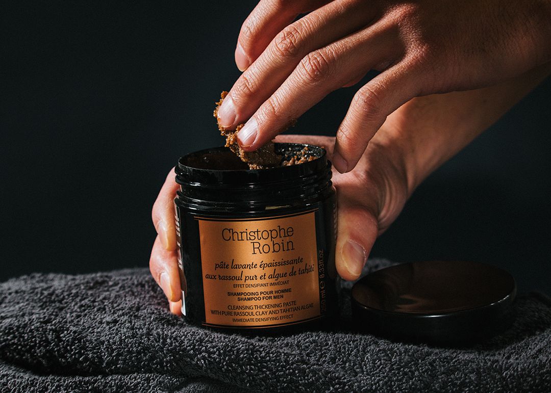 Christophe Robin Cleansing Thickening Paste - Product sits on dark towel in front of a dark background. It's black plastic packaging is glossy with soft light, the label a solid bronze shade. A right hand holds the cylinder packaging while the left hand dips in to take out a teaspoon amount of product. - 1080 × 771