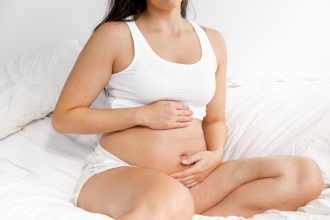 How To Treat Dry and Itchy Skin During Pregnancy - a pregnant woman sitting on a bed with white sheets has her hands on her pregnant belly  1200 x 800