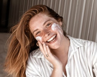 Ultimate Guide to Sunscreen - Woman with red hair and freckles smiling as she applies sunscreen to her face