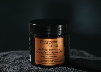 Christophe Robin Cleansing Thickening Paste - Product sits on dark towel in front of a dark background. It's black plastic packaging is glossy with soft light, the label a solid bronze shade. - 1080 × 771