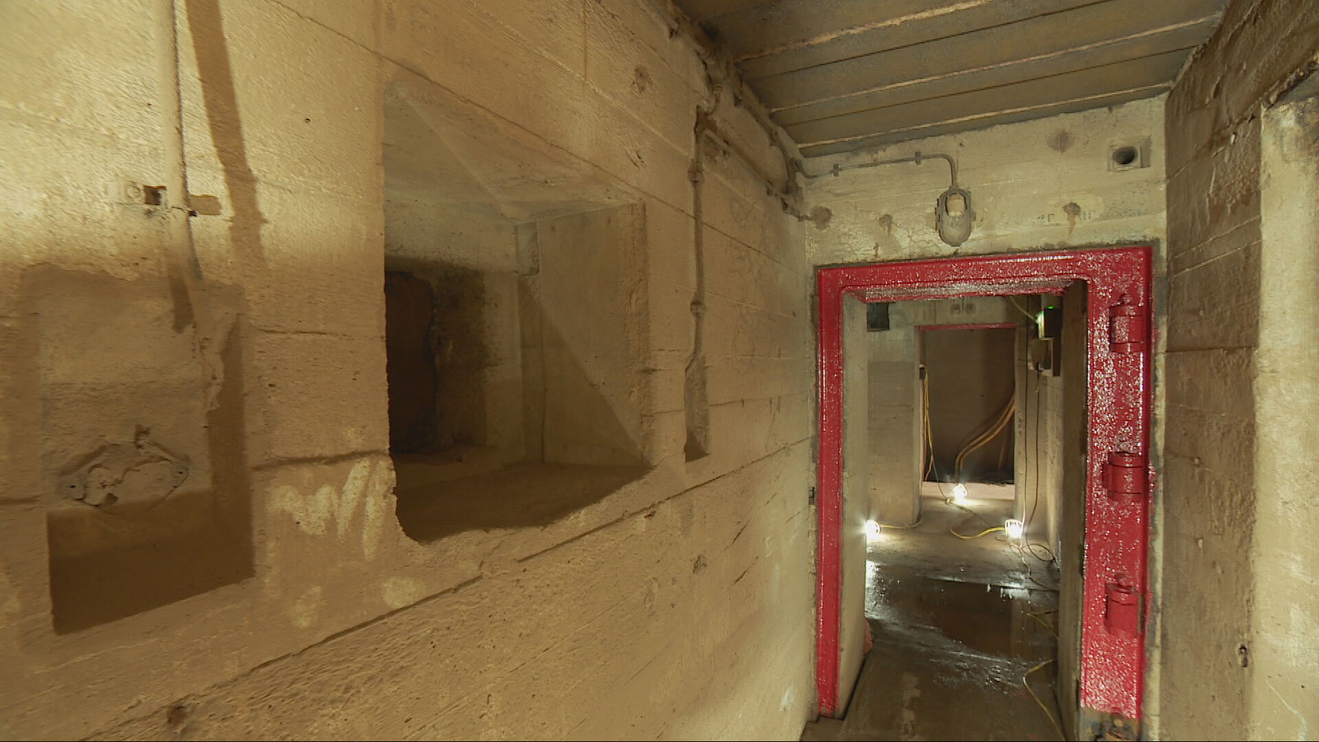An underground passage with a red door frame and floor lights illuminating the stone walls of this underground bunker.