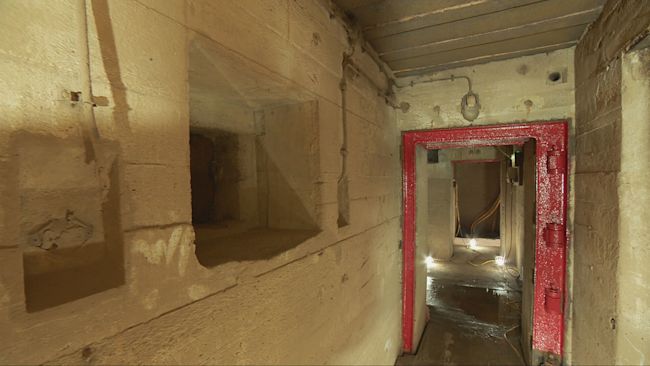 An underground passage with a red door frame and floor lights illuminating the stone walls of this underground bunker.