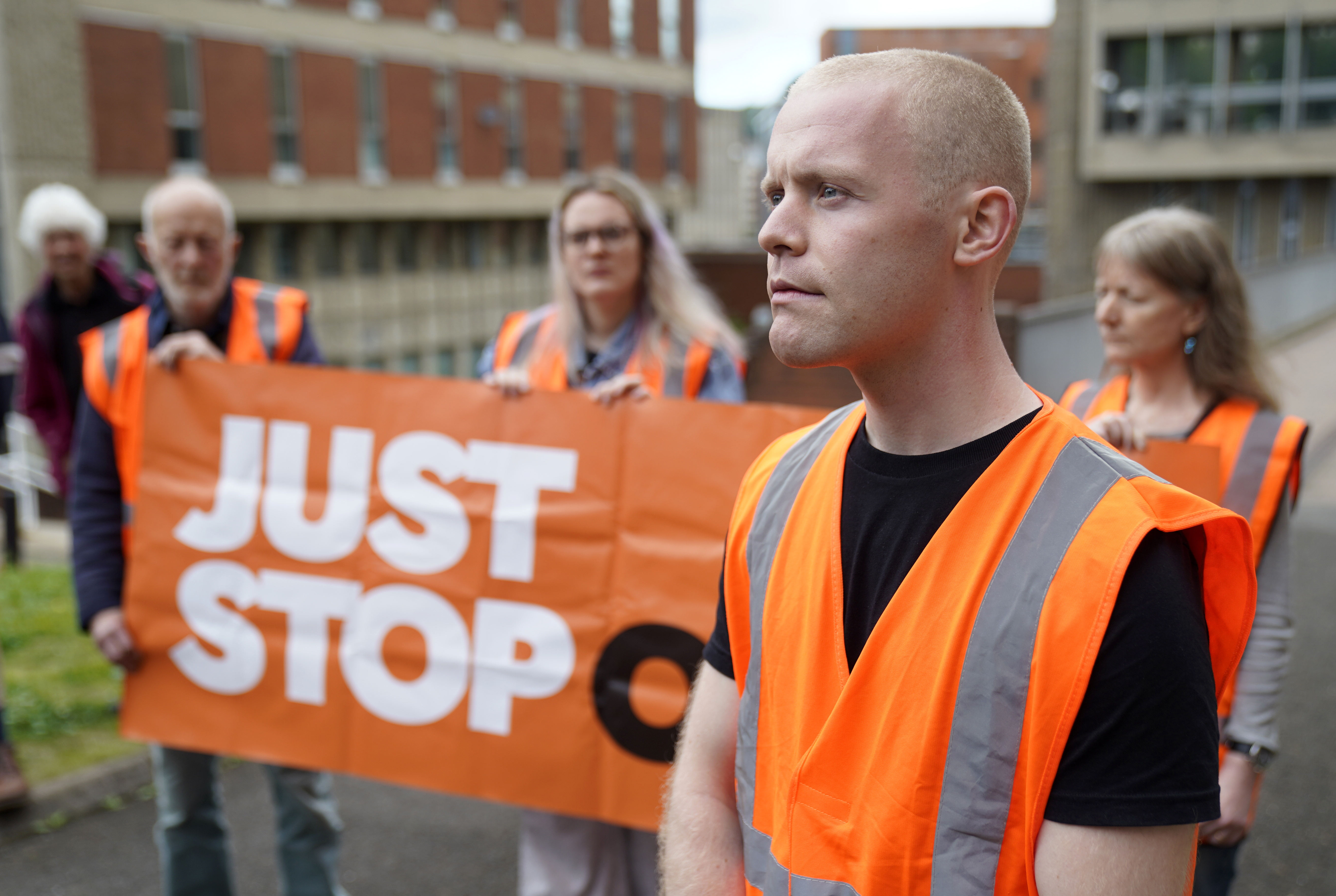 A man in an orange high-visibility vest stands in the foreground outside a brick building. Behind him, supporters hold an orange 