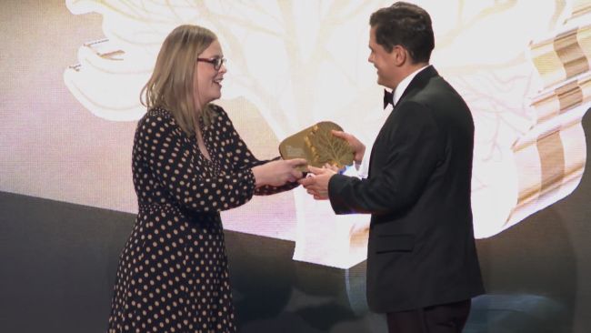 Head teacher of Wymondham school goes to pick up a golden award on the stage as a woman in a spotted dress hands over the award to him. 