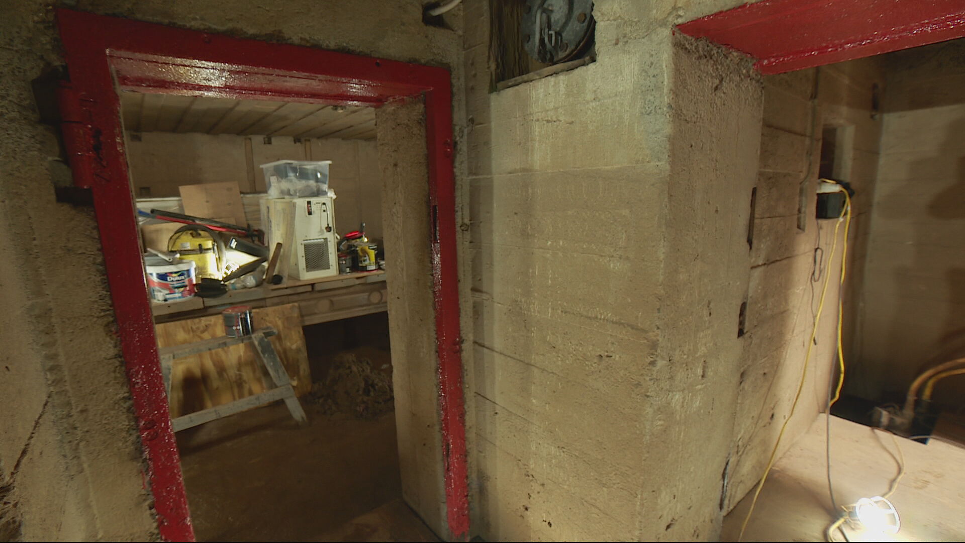 In an underground bunker, a red door frame on the left of the picture leads into a room with a table within with equipment on. A corridor with a light also can be seen on the right of the photo.
