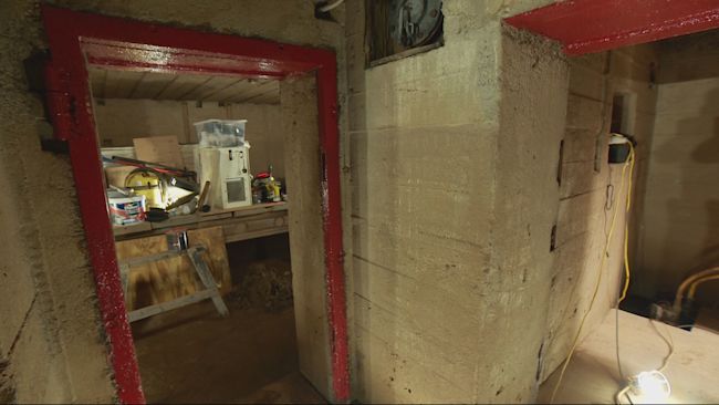 In an underground bunker, a red door frame on the left of the picture leads into a room with a table within with equipment on. A corridor with a light also can be seen on the right of the photo.