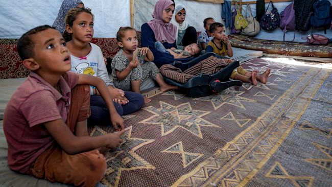 Displaced infant Abdel-Rahman Abu El-Jedian, 11-month-old, who suffers from polio, is carried by his mother, center, at a makeshift tent camp in Deir al-Balah, central Gaza Strip, Tuesday, Aug. 27, 2024. (AP Photo/Abdel Kareem Hana)