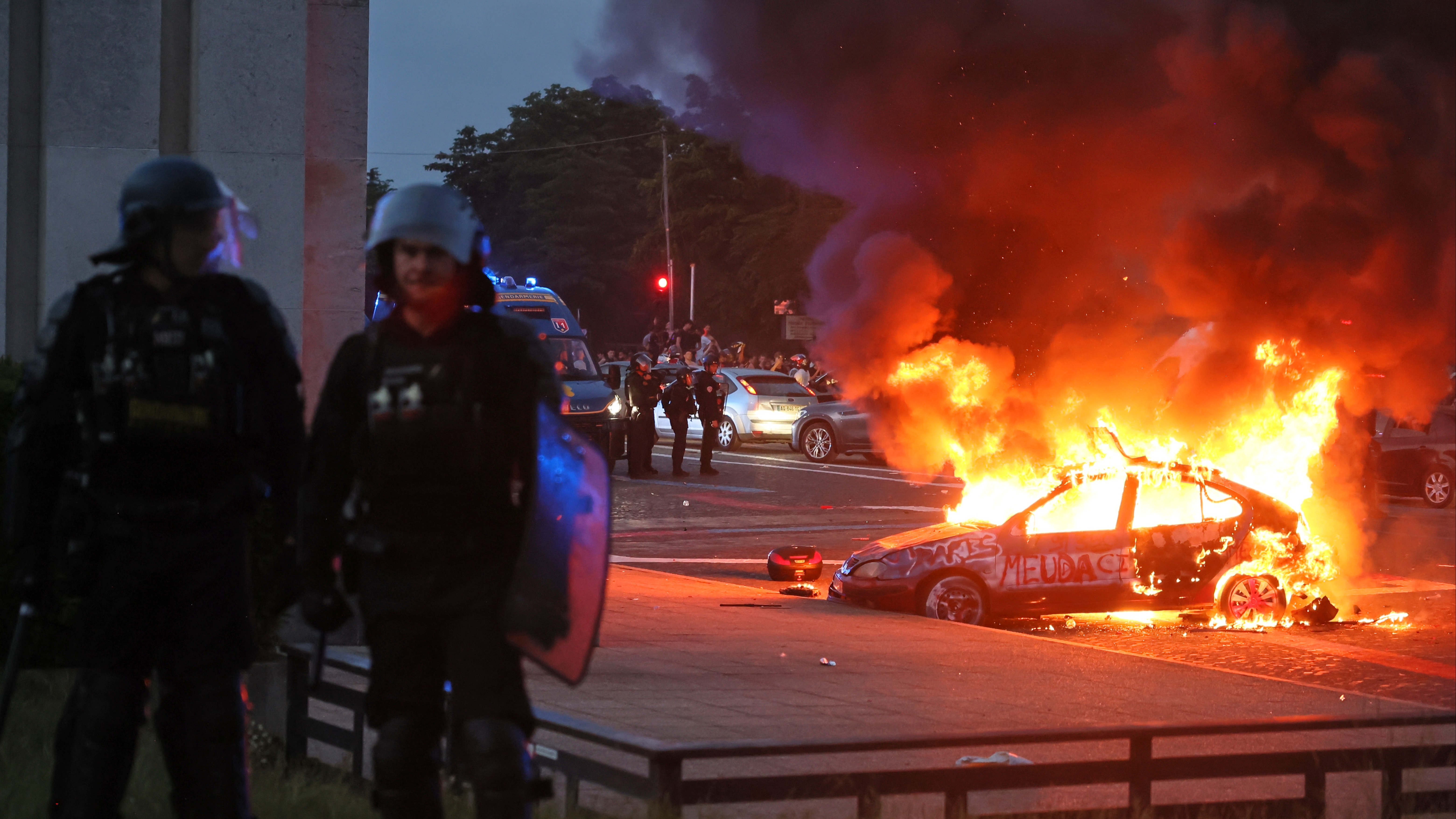 A car ablaze on the streets of Paris with riot police in the foreground.