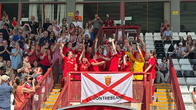 A group of female footballers wearing red kits, jumping and smiling with joy whilst holding a trophy!