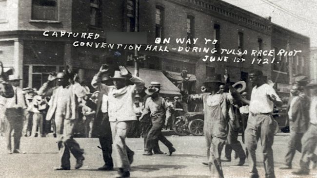 In this photo provided by the Department of Special Collections, McFarlin Library, The University of Tulsa, a group of Black men are marched past the corner of 2nd and Main Streets in Tulsa, Okla., under armed guard during the Tulsa Race Massacre on June 1, 1921.