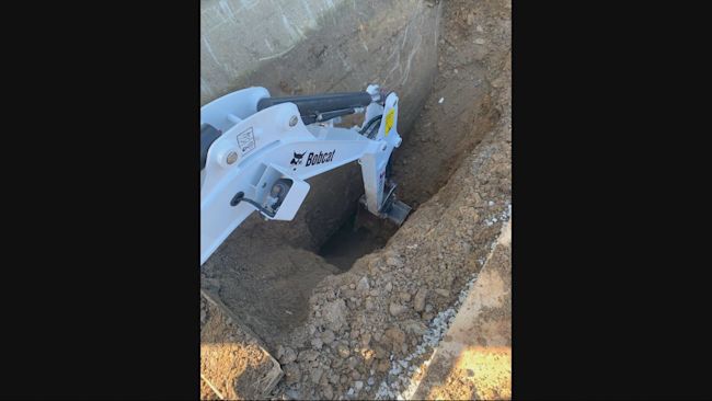 The claw of a Bobcat digger digging a hole with a wall on one side.