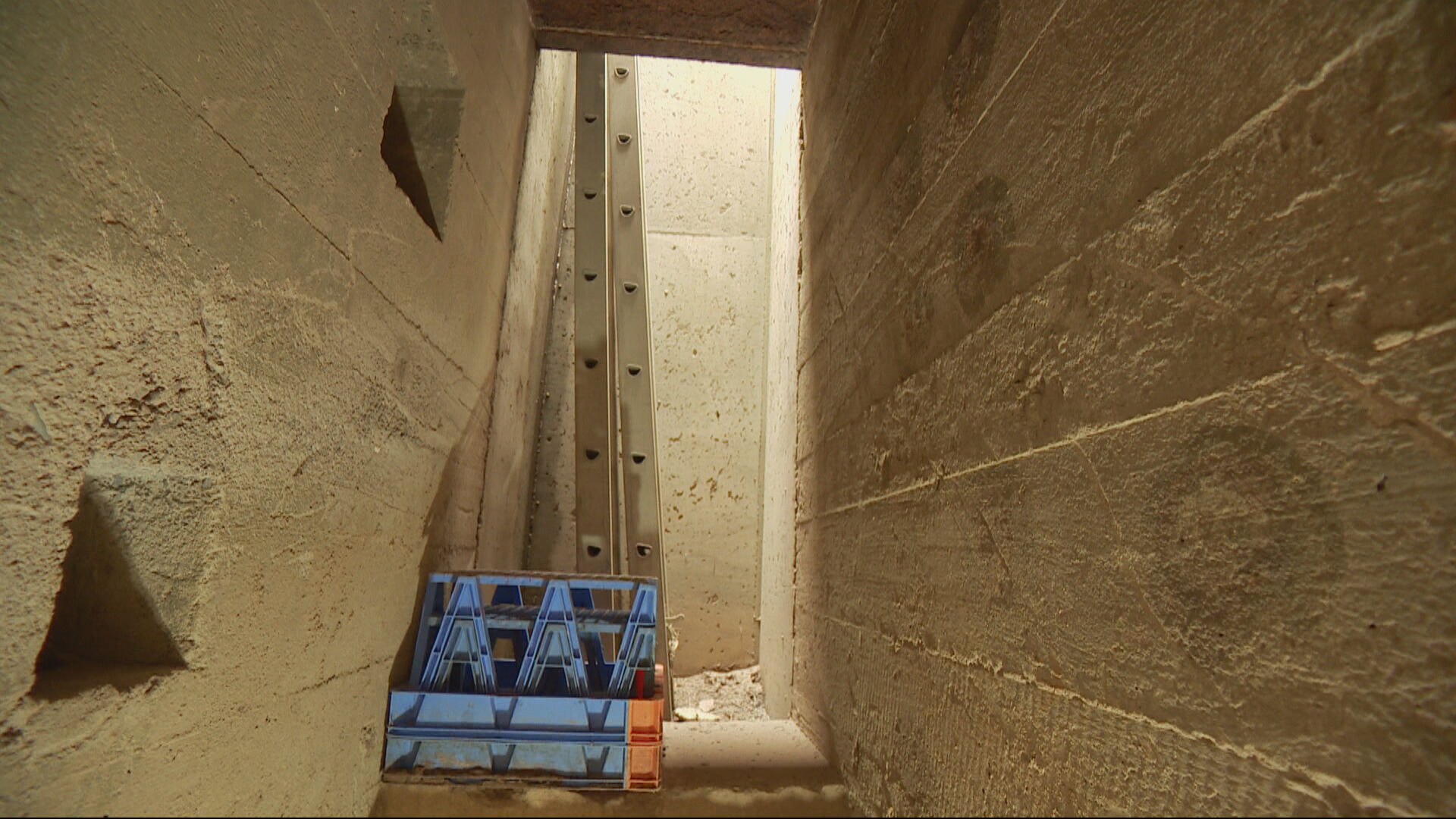 A hatched entrance to an underground corridor with light coming down, with a ladder allowing people to climb down and in.