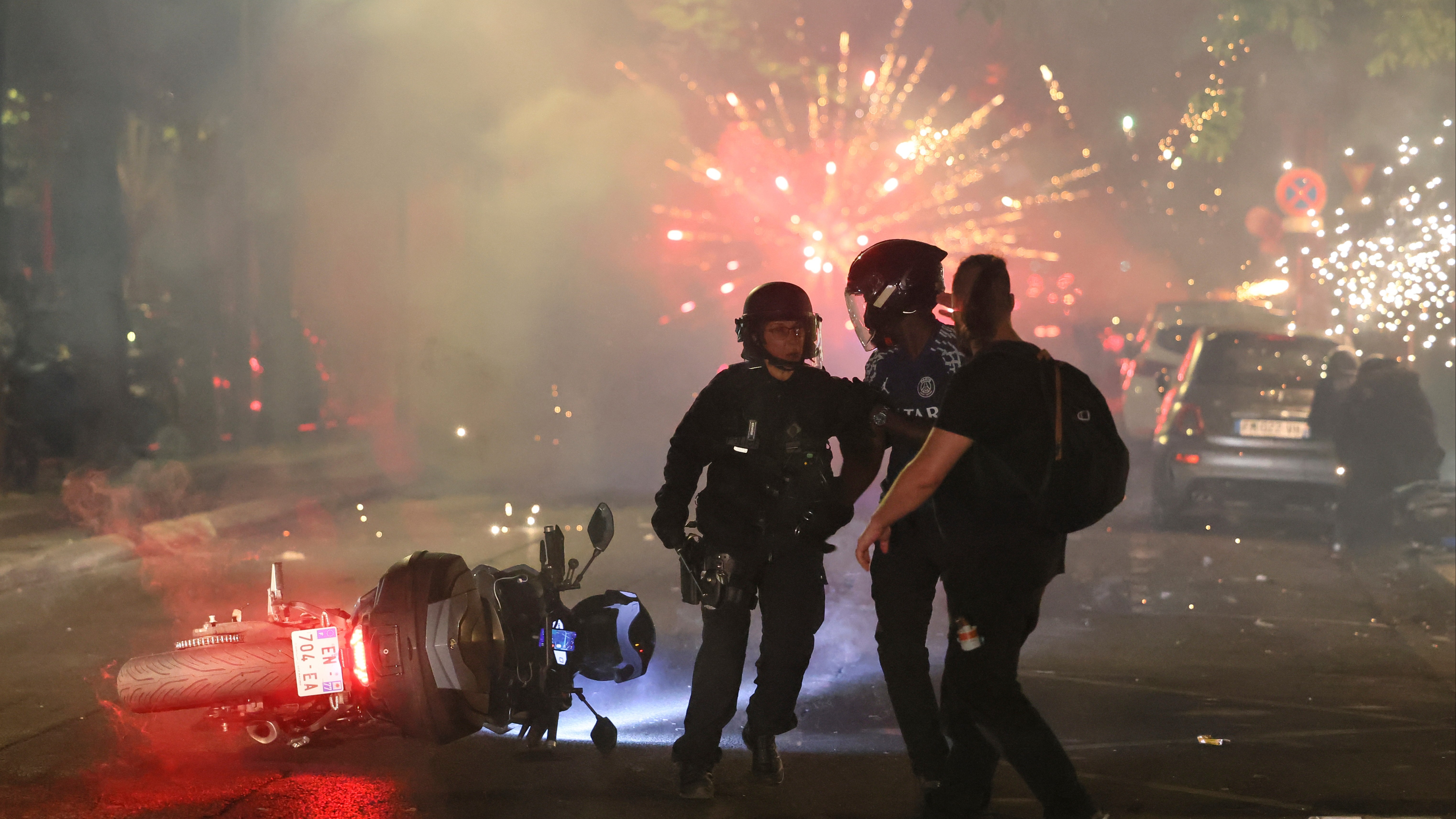 A motorbike lies on the ground by police officers as fireworks explode in the background.