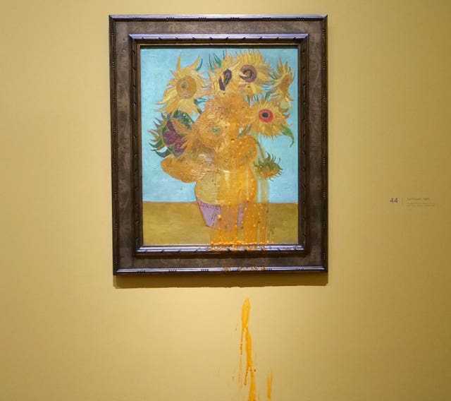 The image shows a framed Van Gogh sunflower painting hanging on a yellow museum wall. The painting depicts vibrant yellow sunflowers in a vase against a blue background. Below the artwork, orange liquid appears to be dripping down the wall, suggesting it has been thrown at the painting. A small museum placard with the number 