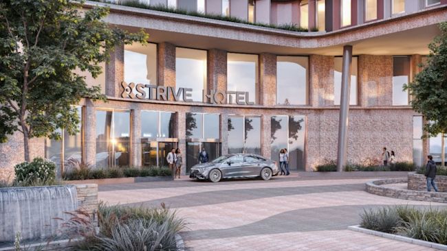 Trees and plants in the foreground with a car pulling up outside the hotel, with a sign that says strive hotel. The building is a mix of glass windows and light granite.