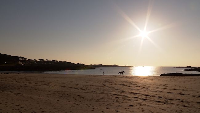 PERSON RIDING HORSE ON BEACH AS SUN BEGINS TO SET IN GUERNSEY