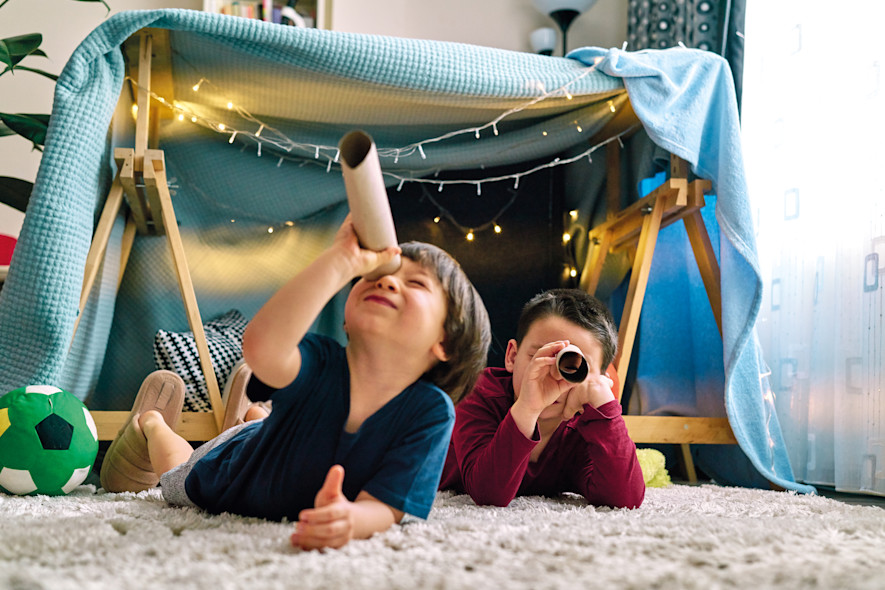 Two children play in the living room in a tent built with tables and blankets