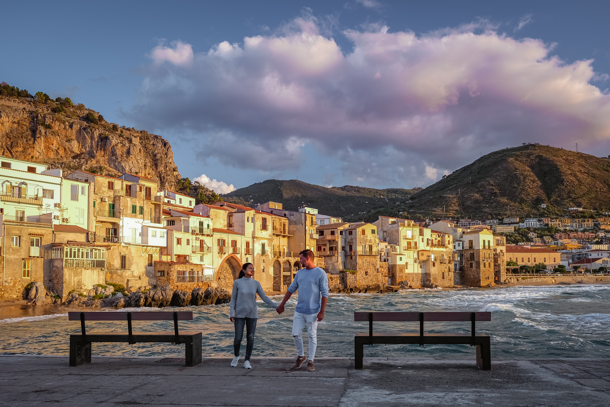 Una coppia si tiene per mano davanti un molo di un paese in Liguria, con il mare mosso sullo sfondo 