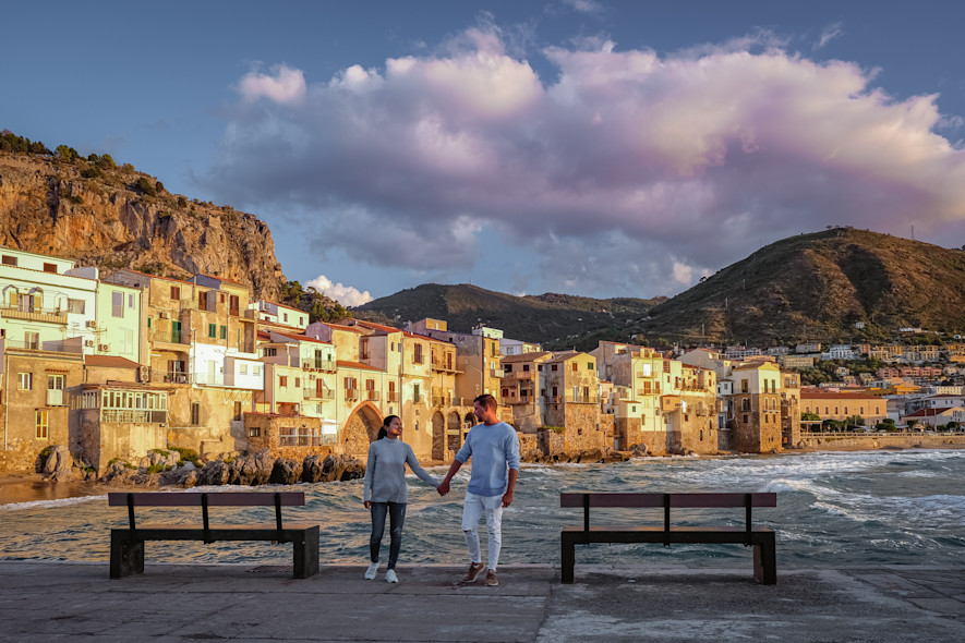 Una coppia si tiene per mano davanti un molo di un paese in Liguria, con il mare mosso sullo sfondo 