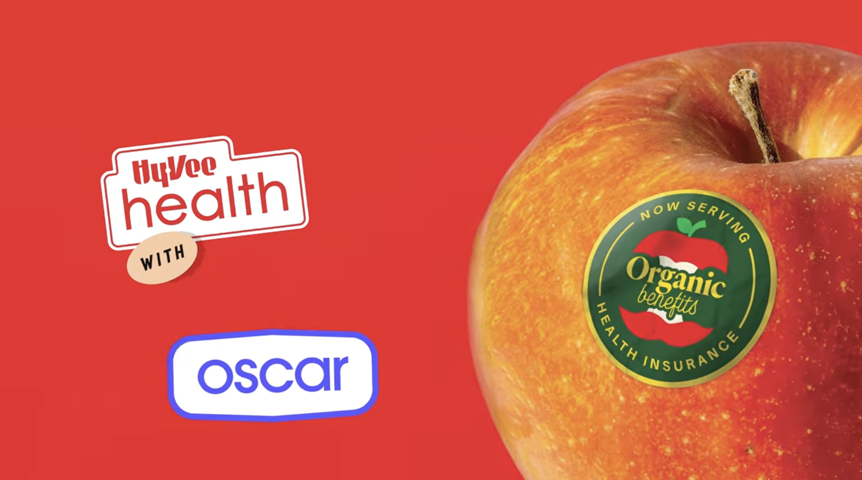 Image of an apple with a sticker that reads "Now serving: Health insurance" and text in the middle that says "Organic benefits". To the left of the apple is the HyVee Health and Oscar logo.