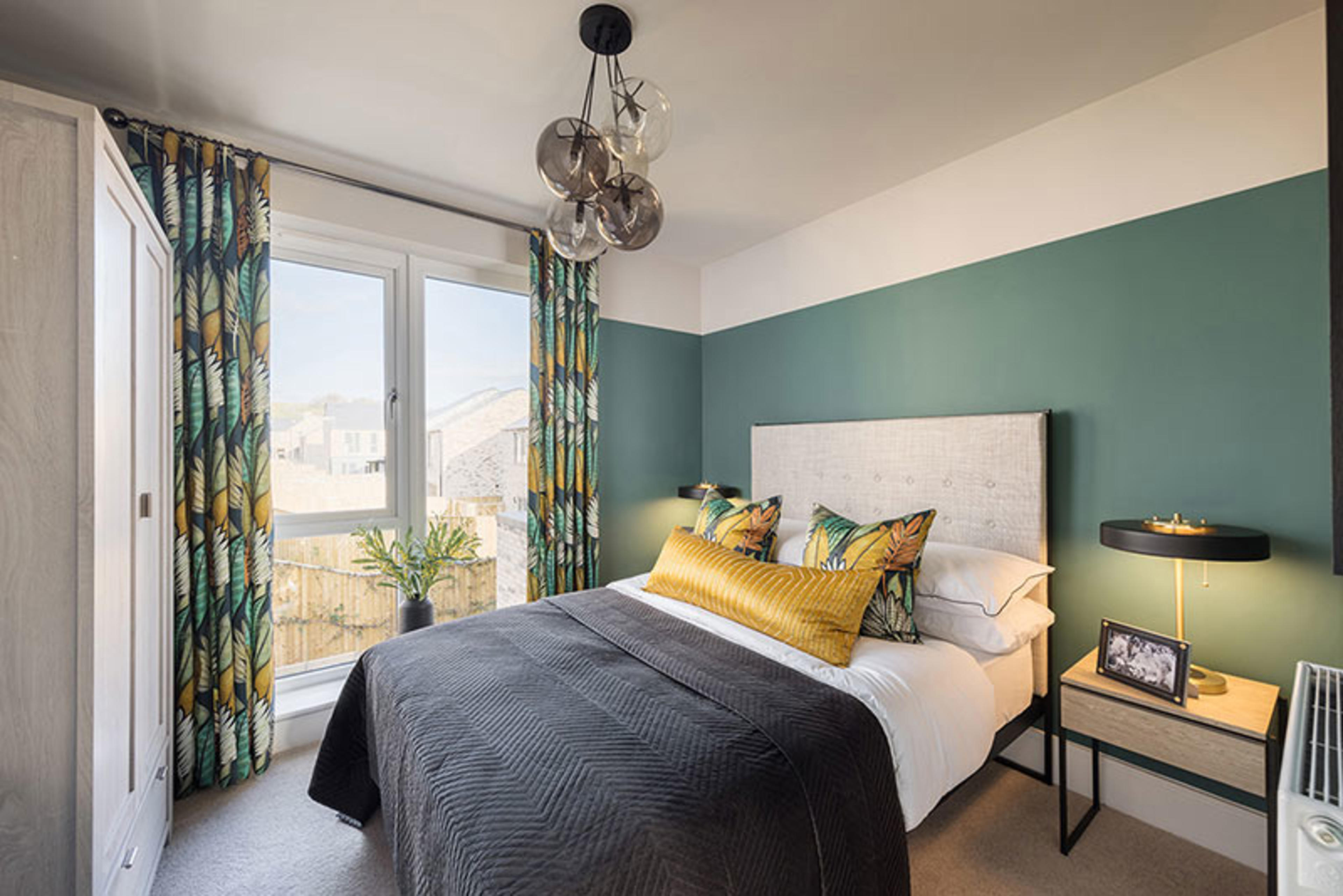 A bedroom with green walls and curtains containing a wardrobe and a double bed with green and yellow pillows 