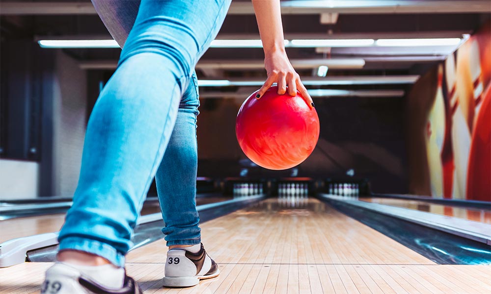 A woman in blue jeans ten-pin bowling with a red bowling ball 