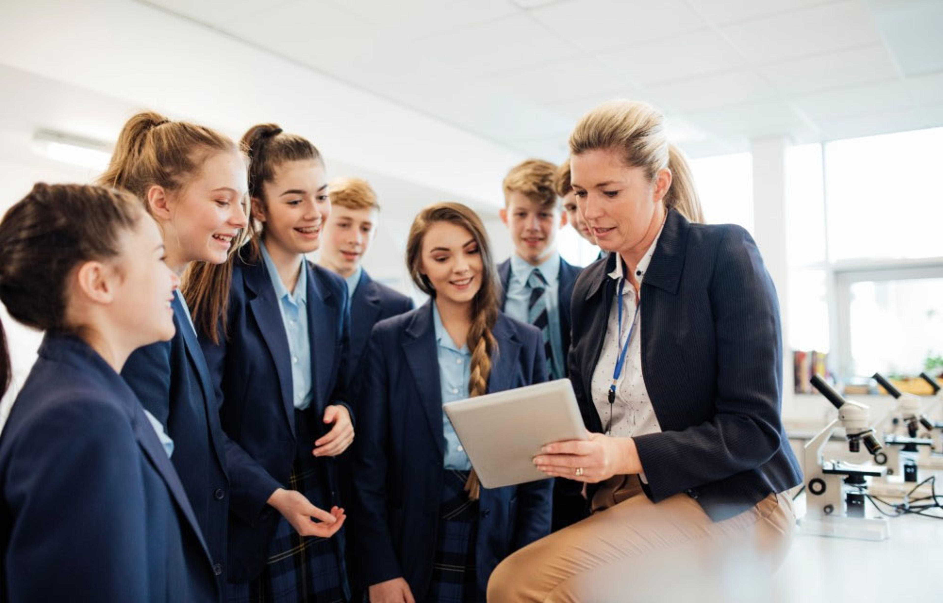 A teacher showing a tablet to a group of secondary school pupils in a science classroom, microscopes visible on a table.