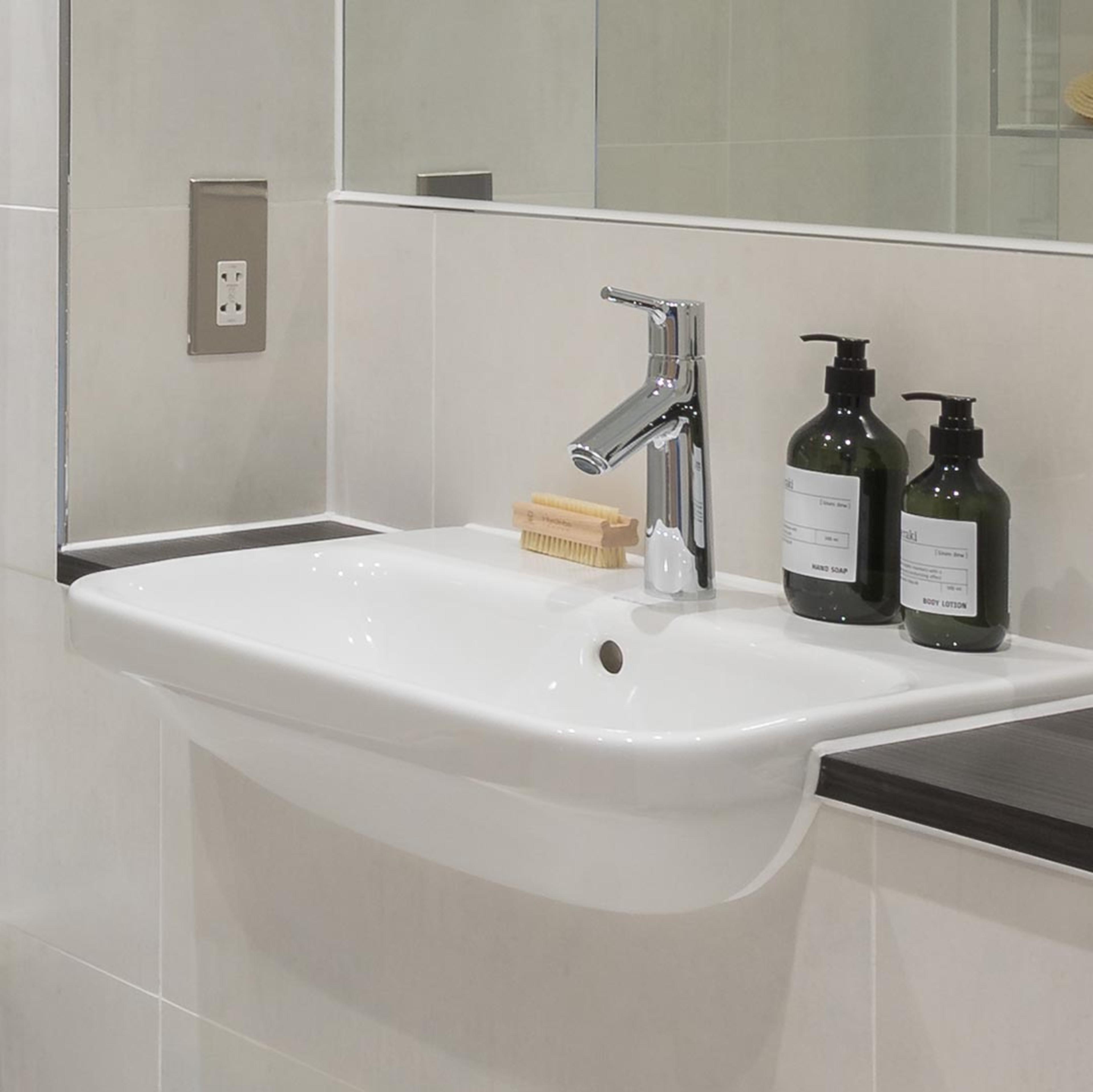 A white sink with chrome mixer tap in a cream tiled bathroom with a mirror behind. Bottles of facewash/handwash stand on the sink.