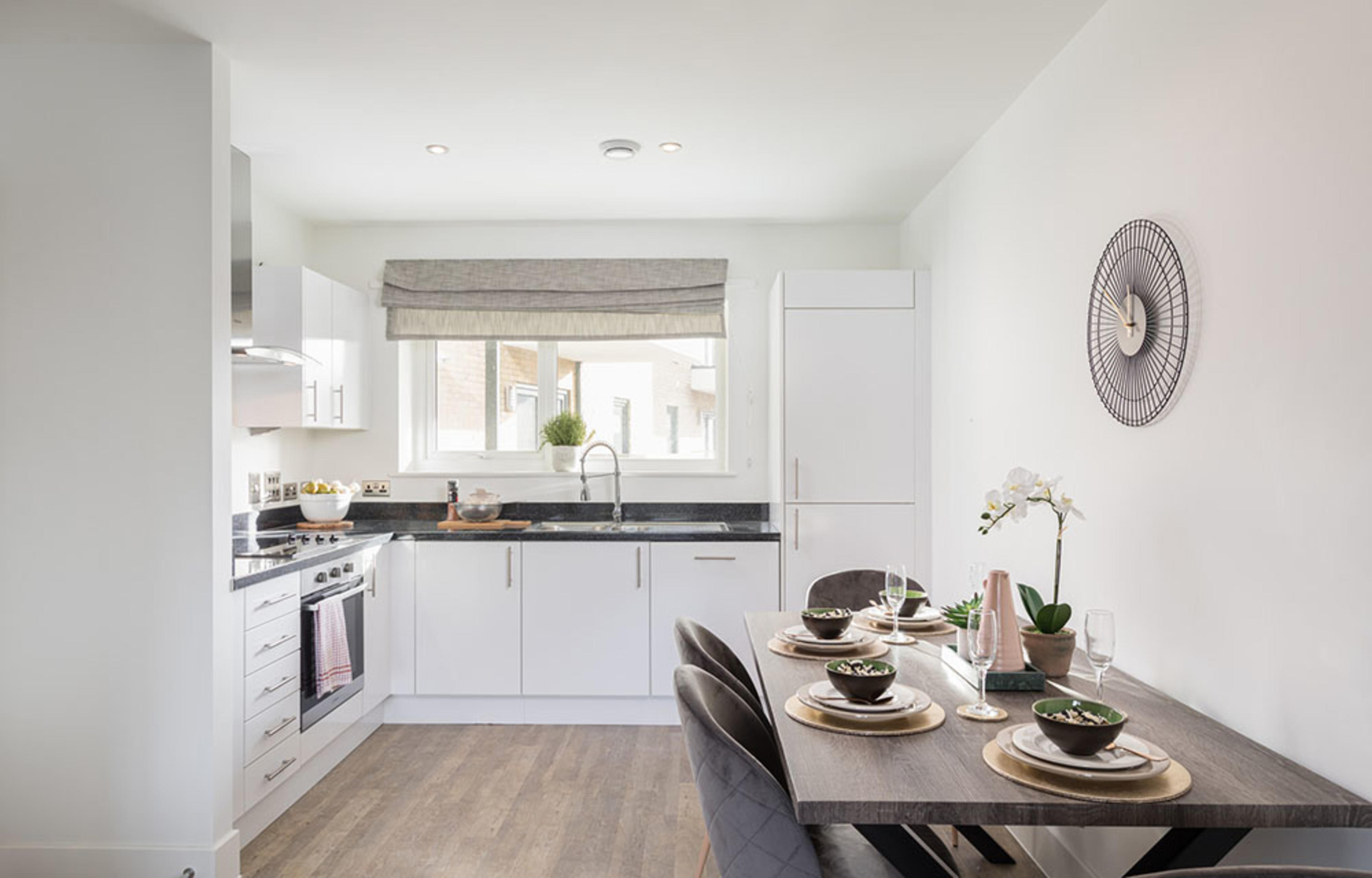 An L-shaped kitchen with white cabinets, dark worktops and up-stands and brushed silver appliances. The dining area contains a rectangular stone-effect table set against the wall.