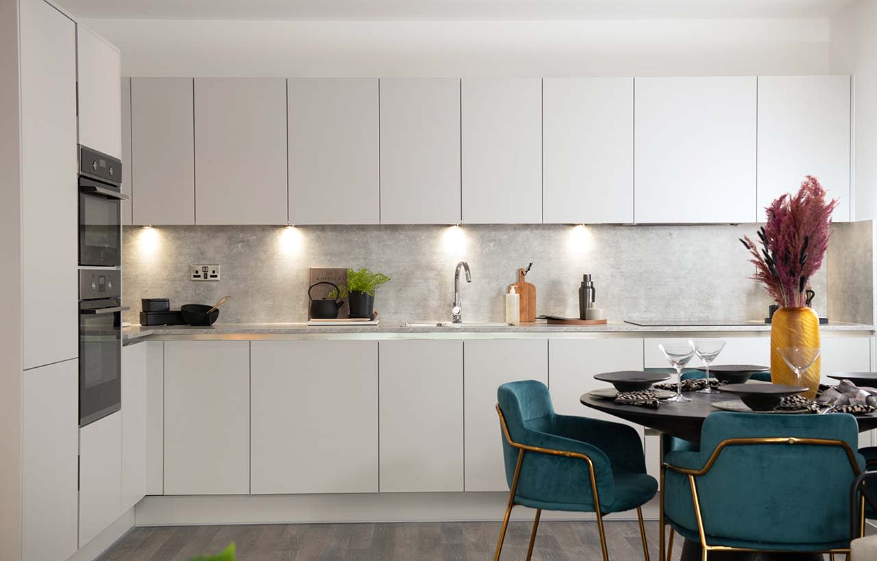 CGI of apartment kitchen/dining area with handleless slab kitchen units in white, double oven, and dining table with green and gold chairs