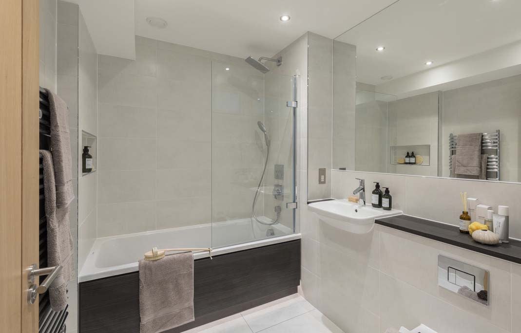 Fully-tiled bathroom in pale cream with wall-width mirror, bath with dark wood-effect panelling and ceramic tiled floor.