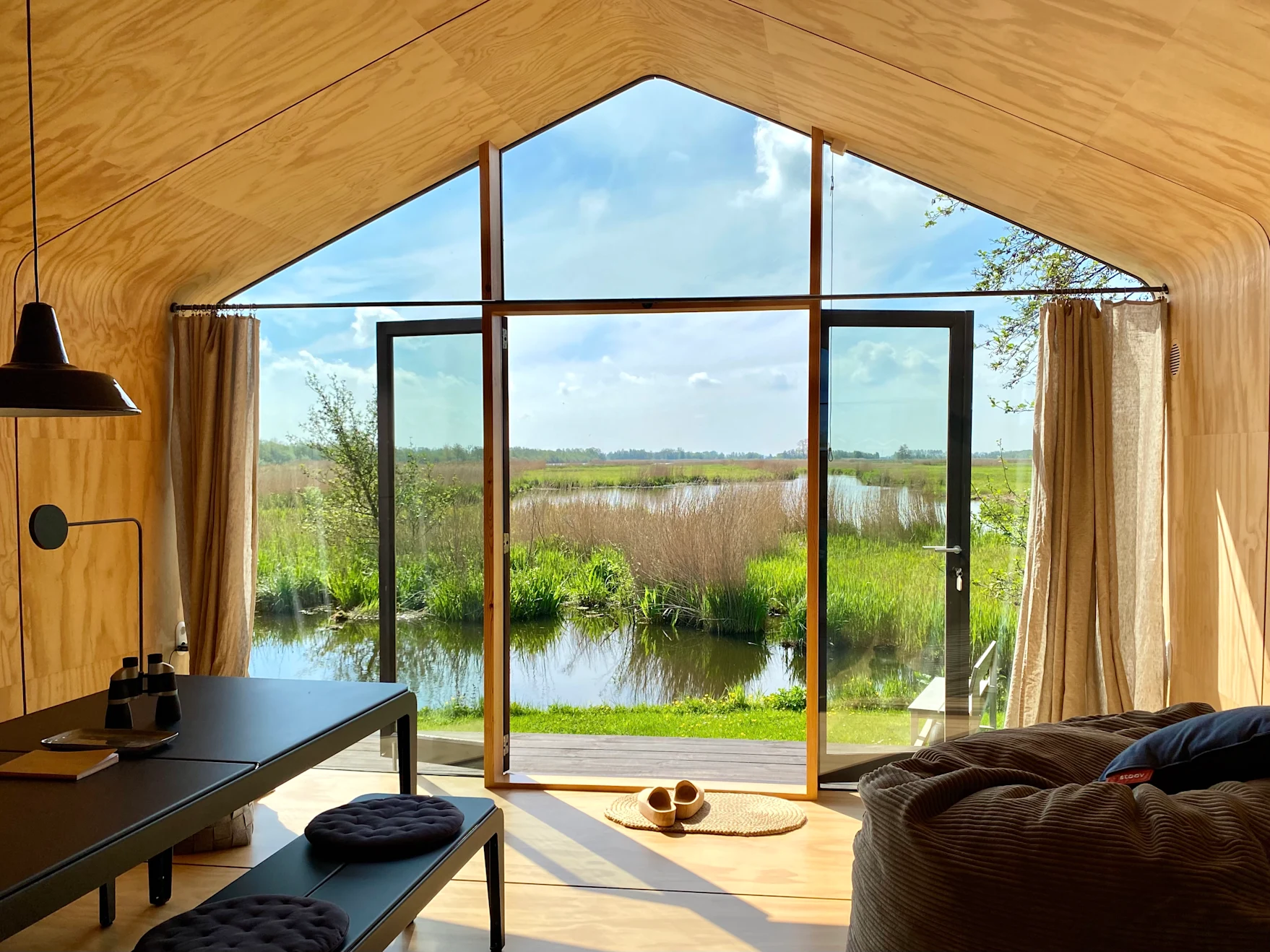 Vue depuis l'intérieur d'une maison en bois, avec vue sur un lac. Le soleil inonde la pièce à travers les portes vitrées ouvertes.