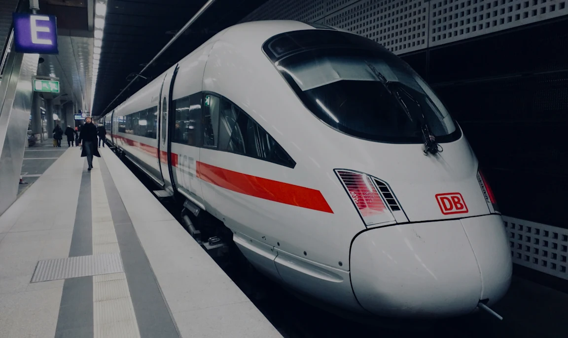 L’image montre un train à grande vitesse à quai dans une gare. Le train, au design élégant, est blanc avec une bande rouge et le logo « DB » sur l’avant. 