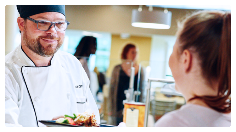 A man in a chef's uniform hands a plate of food to a woman at a buffet.