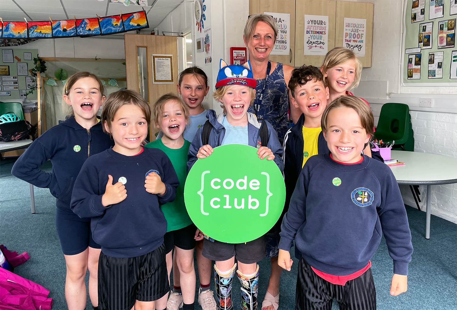 A group of young Code Club learners and their teacher stand together holding a Code Club sign