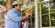 A man in a baseball cap stands on a ladder and applies caulking to a large house window.