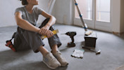 A woman holding pencils sits next to a ladder and some tools in an unfinished room