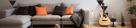 A gray couch with black and orange pillows sits next to a guitar and house plants.