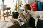 Woman on laptop sitting on the floor of her apartment, white dog lounges on the couch behind her and rests its head on her shoulder.