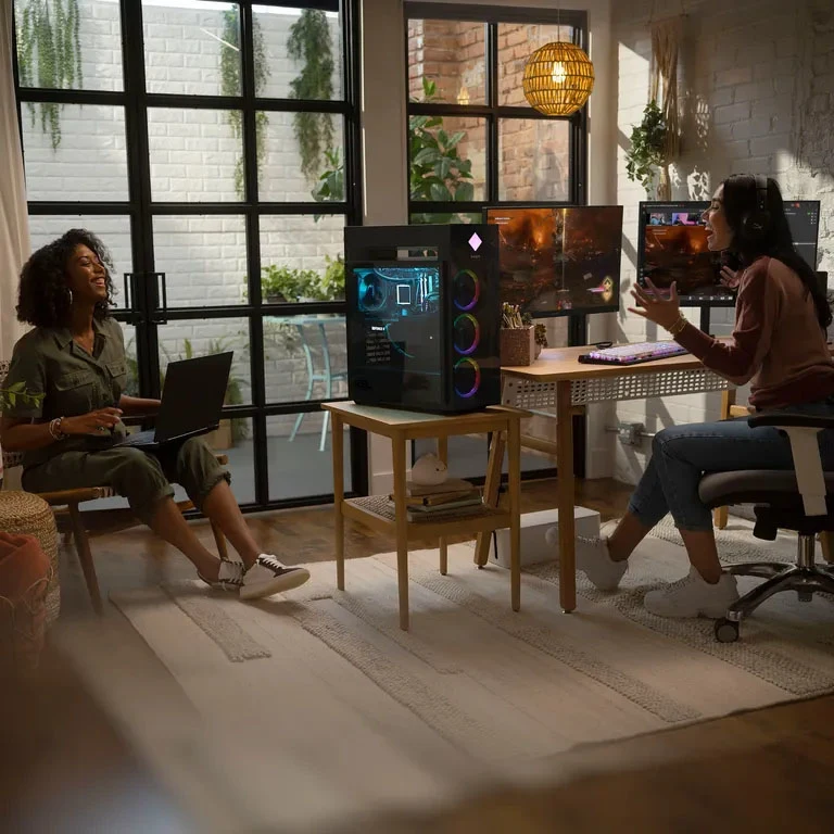 Two women sitting in a room with a laptop and a fixed PC.