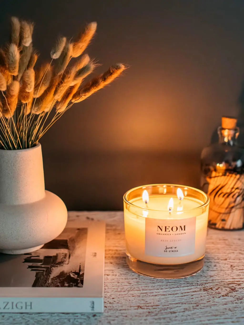 A Neom candle lit and placed on a shelf with a book, matches and dried flowers