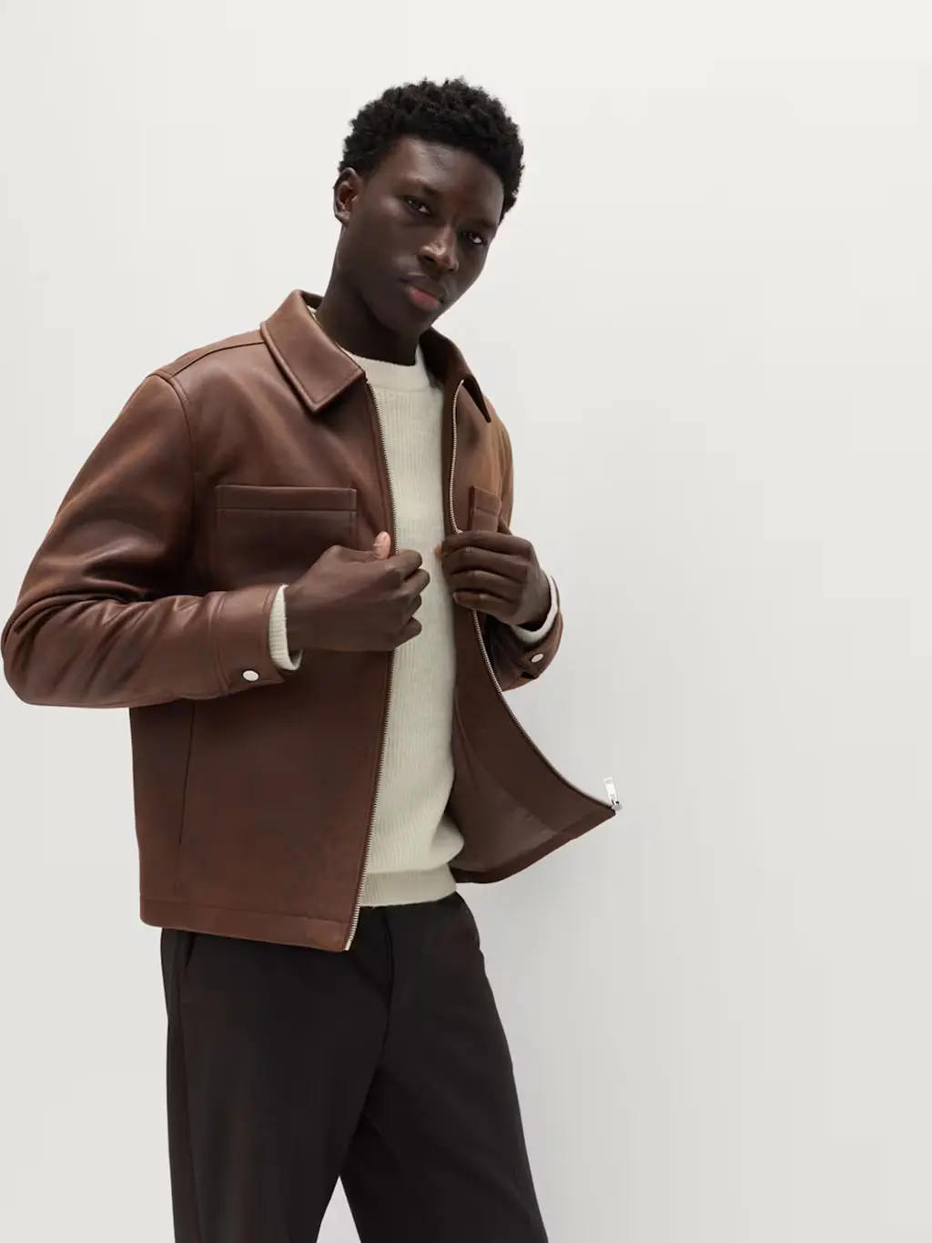 Man wearing a cream jumper with brown tailored trousers and a brown leather jacket