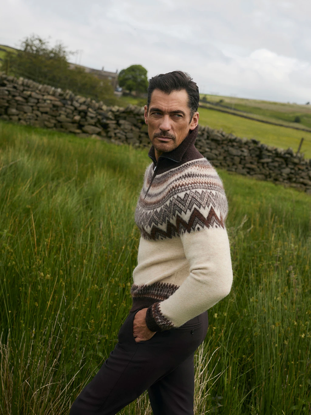 Man wearing a fair isle knitted jumper and brown trousers, walking through a field.