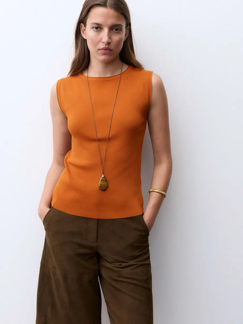 Woman wearing a rust-toned top with brown trousers and a long necklace