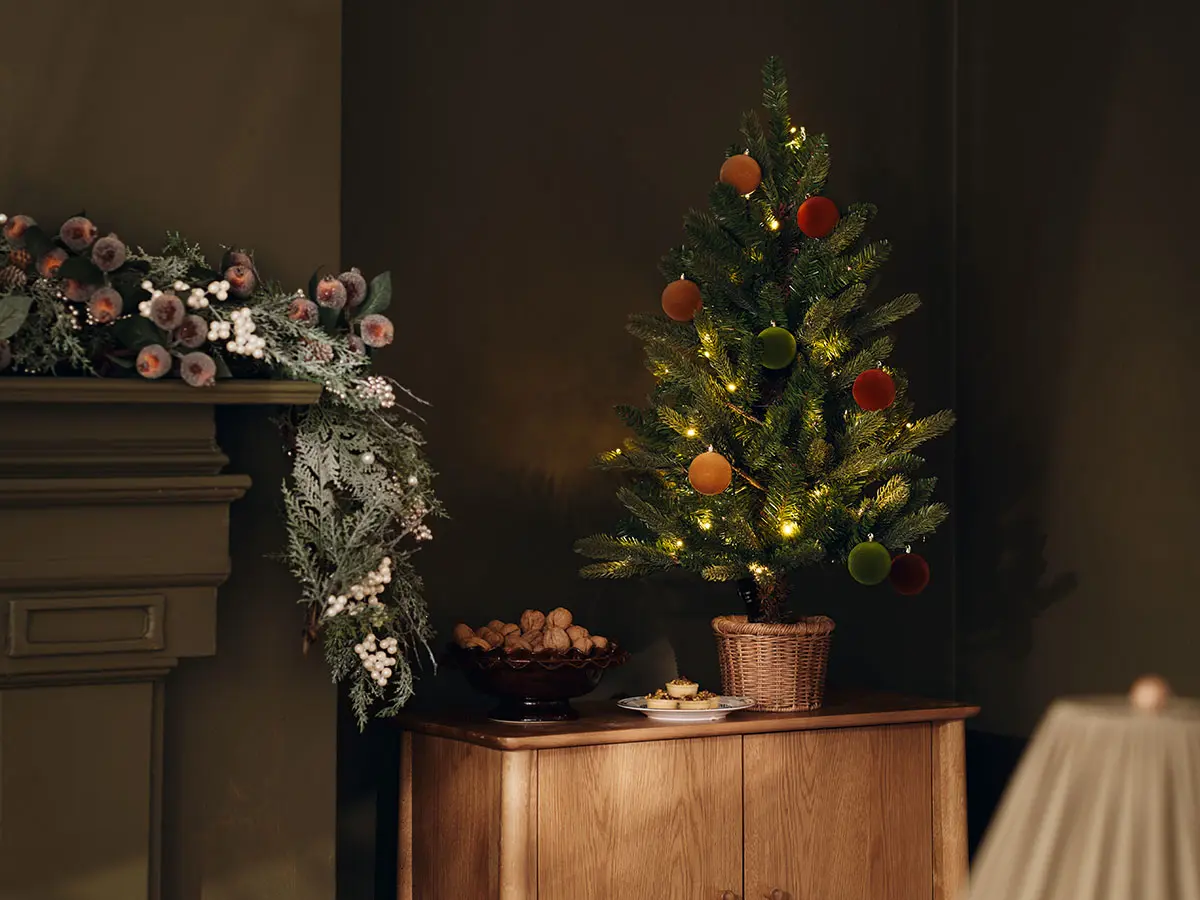 Small dressed Christmas tree on side cabinet next to mantelpiece covered in festive foliage