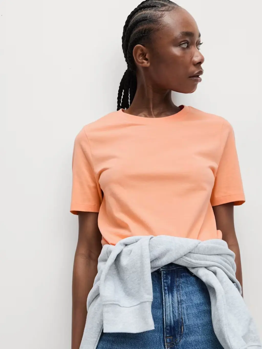 A woman wears a pale orange T-shirt with jeans and a grey sweatshirt tied around her waist