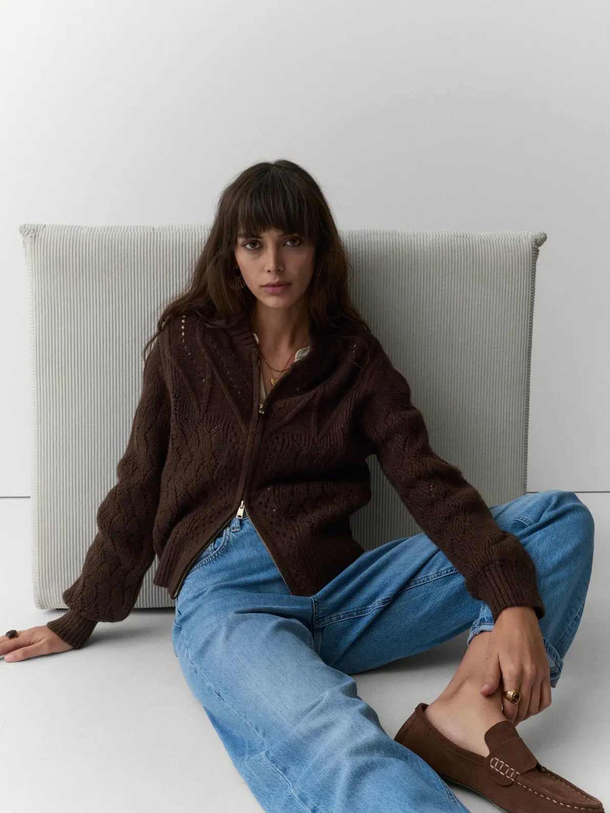 Woman sitting on floor, wearing brown zip-up cardigan, blue jeans and brown suede loafers