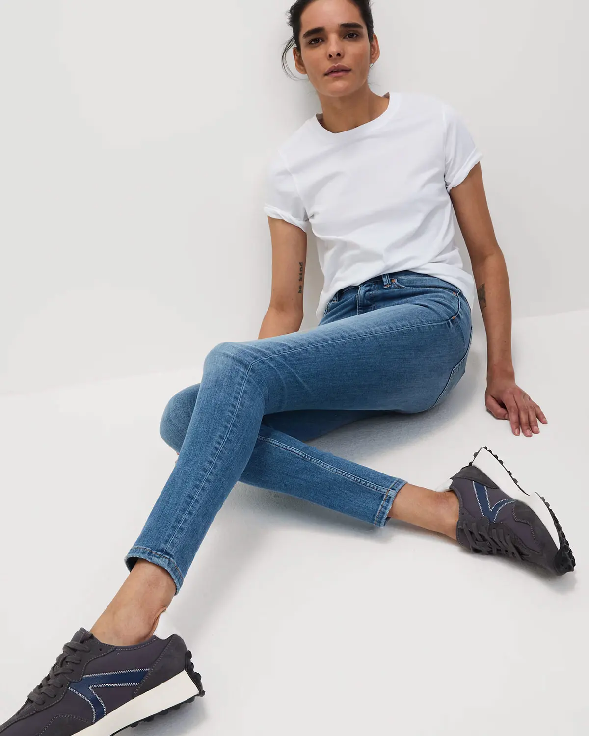 Woman sitting down wearing white T-shirt, blue skinny jeans and black trainers